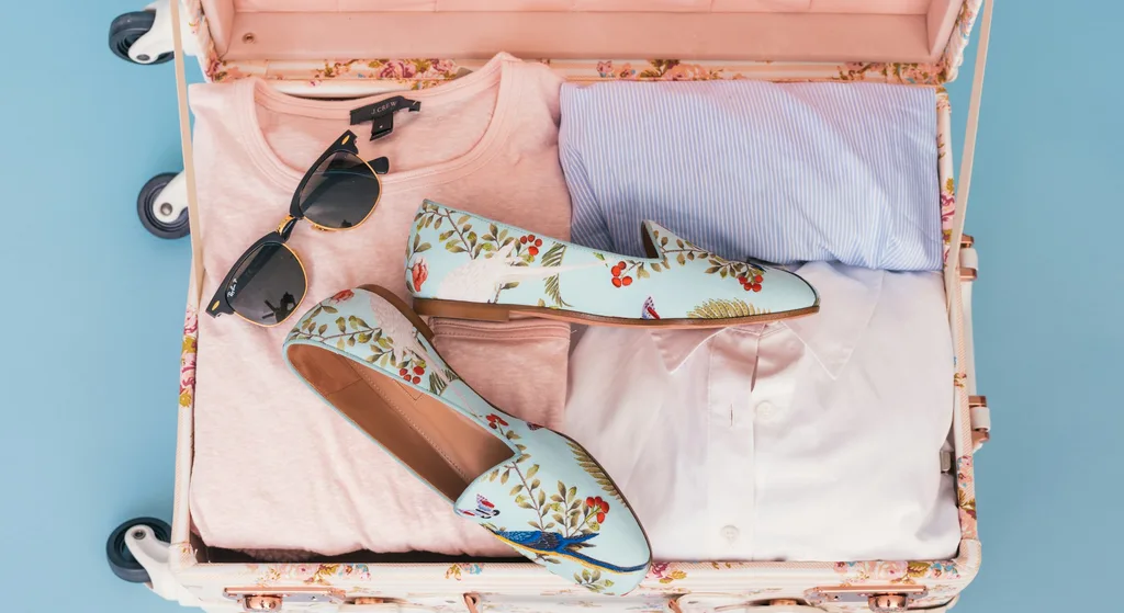 An open suitcase packed with folded clothes, sunglasses and a pair of patterned flats